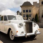 Vintage white Rolls-Royce parked in front of a grand mansion.