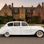 White vintage car parked in front of a stone manor.